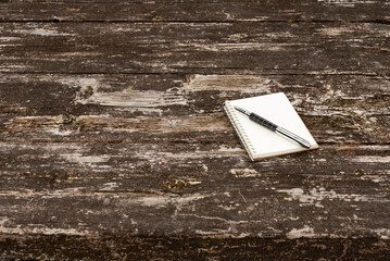 Minimalist scene: Notepad with metal quill pen on rustic wood table, positioned right. Top view in natural daylight. Significant copy space left. Symbolizes planning and writing.