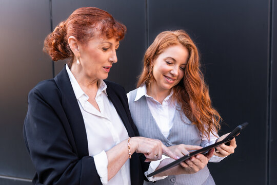 Mature Businesswoman mentoring adult daughter discussing work on a digital tablet outside office - Powered by Adobe