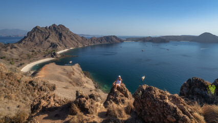 A breathtaking vista showcasing Padar Island, woman sitting on a rock, tranquil bay that features rocky terrain, vibrant boats, and shimmering clear blue waters beneath sunny sky, Komodo, Indonesia