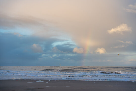 A faint rainbow glows through pastel clouds over the rolling sea. Gentle waves meet the quiet shore in soft evening light.