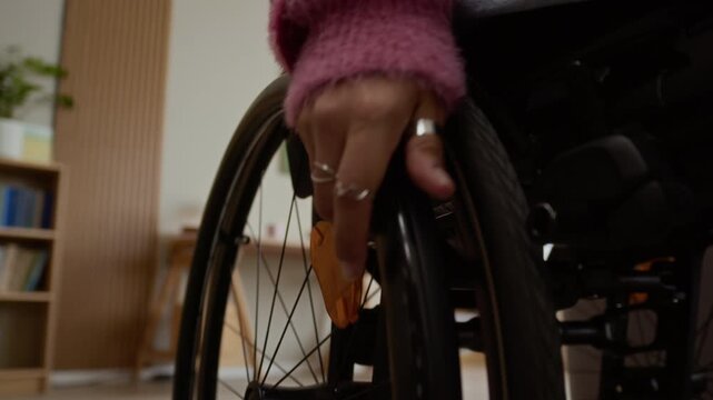 Cropped POV shot of hand of unrecognizable woman with disability pushing wheel forward riding in wheelchair in living room while spending time at home
