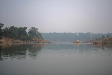 calm stretch of the narmada river surrounded by rocky banks and greenery, with a distant bridge visible through soft mist