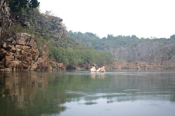 calm river flows between rugged canyon cliffs and lush greenery, with exposed marble rocks emerging from the water, capturing the serene untouched beauty of nature near bhedaghat, jabalpur