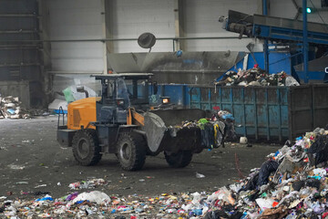A large machine moves through a waste sorting facility, handling mixed household garbage and preparing materials for recycling. Emissions and waste are evident within the industrial space