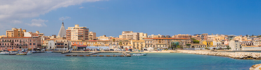 Panoramic Ortigia waterfront in Syracuse, Sicily&mdash;pastel historic buildings, marina and boats behind a stone breakwater under a clear Mediterranean sky; serene coastal cityscape.