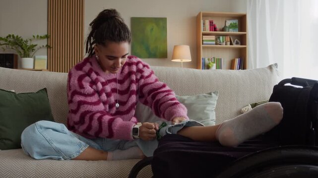 Full shot of motivated young Caucasian girl with disability sitting on couch in cozy living room and using triangle ball massager on legs placed on wheelchair nearby for better recovery effect