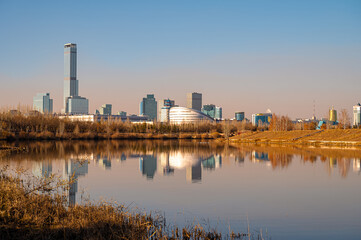 Obraz premium Autumn cityscape of Astana with Abu Dhabi Plaza, modern buildings and river reflection under clear blue sky