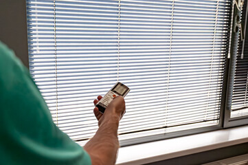 Hand holding remote control to operate modern horizontal blinds on window. Concept of smart home,...