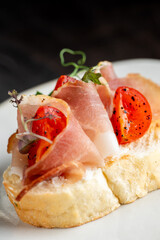 Appetizer bruschetta with thinly sliced serrano ham, roasted cherry tomatoes, fresh herbs, and olive oil on a piece of toasted bread, shot with a shallow depth of field