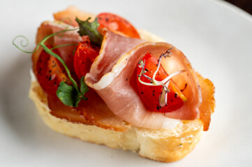 Appetizer bruschetta with thinly sliced serrano ham, roasted cherry tomatoes, fresh herbs, and olive oil on a piece of toasted bread, shot with a shallow depth of field