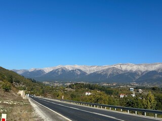 Naklejka premium A clear autumn day unfolds beneath a vast blue sky, as a winding road leads toward majestic snow-dusted mountains and a peaceful valley town surrounded by green forests and golden trees.