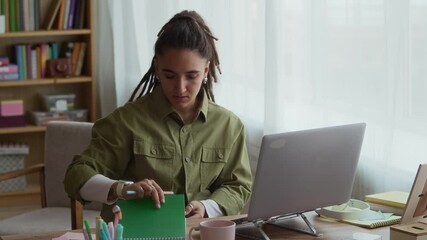 Medium shot of focused young Caucasian girl with disability sitting in front of laptop at table in cozy living room and opening notebook getting ready to study online or work from home