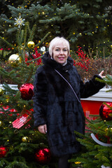 A smiling white woman with short hair stands by a decorated Christmas tree. She's wearing a black...