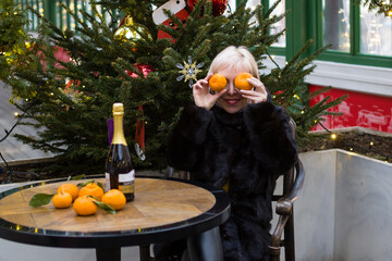A woman with short blonde hair is sitting at a table. She holds a drink in her hands and smiles....