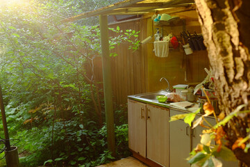 Outdoor kitchen sink area surrounded by lush green plants and trees, sunlight streaming through foliage, stainless steel sink with dishes and utensils, rustic wooden fence in background