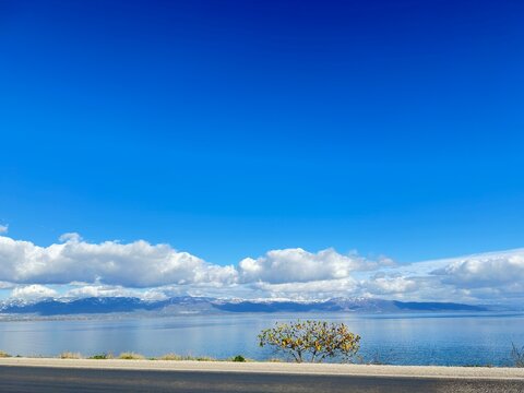 Calm waters mirror the snow-dusted mountains and drifting clouds beneath a brilliant blue sky, capturing the peaceful harmony of nature where earth, water, and sky unite in quiet perfection.