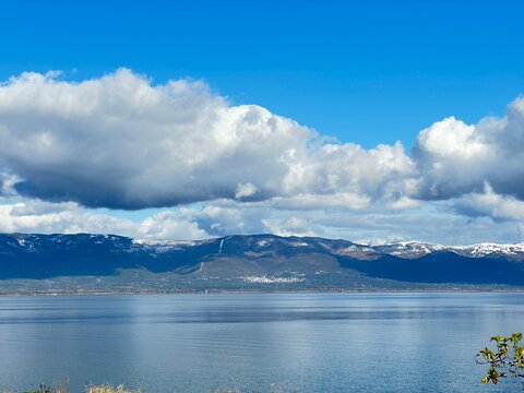 Calm waters mirror the snow-dusted mountains and drifting clouds beneath a brilliant blue sky, capturing the peaceful harmony of nature where earth, water, and sky unite in quiet perfection.