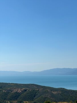 A serene panoramic view of a turquoise lake surrounded by lush green hills and distant blue mountains under a clear summer sky, capturing the calm beauty of untouched nature in perfect harmony.