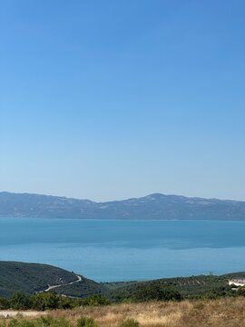 A serene panoramic view of a turquoise lake surrounded by lush green hills and distant blue mountains under a clear summer sky, capturing the calm beauty of untouched nature in perfect harmony.