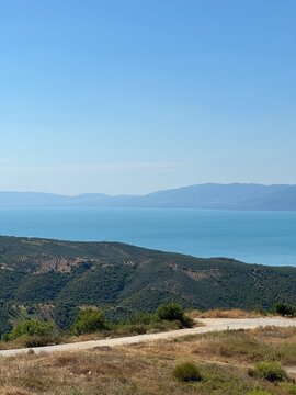 A serene panoramic view of a turquoise lake surrounded by lush green hills and distant blue mountains under a clear summer sky, capturing the calm beauty of untouched nature in perfect harmony.