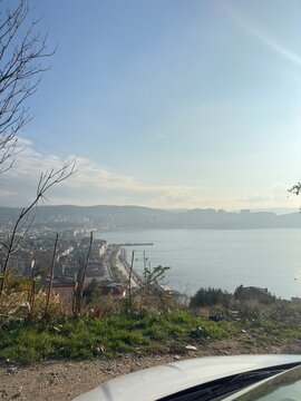 A stunning coastal city view from a hillside overlooking calm blue waters, sunlit buildings, and a peaceful shoreline stretching into the horizon under a bright, clear sky on a serene afternoon.