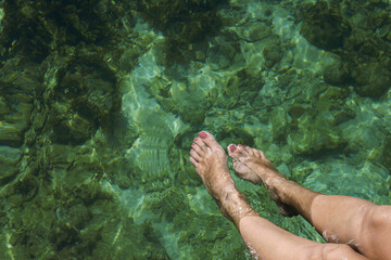 Caucasian young adult woman relaxing with legs submerged in clear water, sunlight creating patterns on skin and surface, natural outdoor setting, only lower legs and feet visible