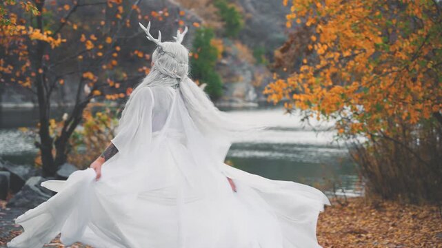 fantasy happy elf woman beauty face dancing spinning in autumn nature yellow trees. sexy adult fairy tale girl long blonde ash hair white dress snow queen style makeup. myth faun deer antlers on head