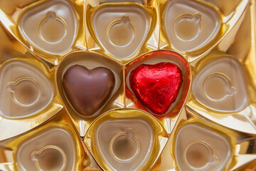 Two heart-shaped chocolate candies in a golden box, one candy in a red wrapper and the other unwrapped, close-up.