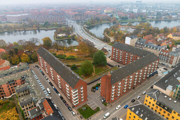 Copenhagen - An aerial view captures V-shaped brick residential buildings, a road with traffic, and a bridge crossing a lake system surrounded by autumn trees under an overcast sky.