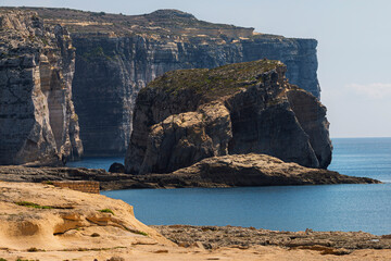 Malta - Dramatic sheer cliffs and a large sea stack rise from the turquoise water under a clear sky along a rugged coastline.