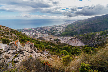 Fototapeta premium Spain - From a rocky mountain vantage point, a coastal city and highway stretch along the ocean under a dramatic cloudy sky.