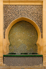Old Moroccan wall fountain in Fes medina covered in traditional zellige tiles and geometric Islamic motifs, dry basin below, showcasing intricate craft and cultural identity.