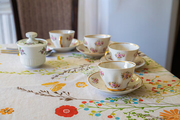 A collection of floral teacups and a sugar bowl are elegantly arranged on a patterned tablecloth, ready for a gathering.