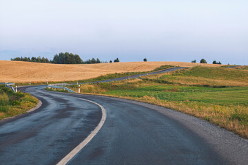 Fototapeta premium Lithuania - A curving asphalt road winds through golden wheat fields and green grass under a clear sky in a peaceful rural landscape.