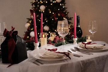 A festive holiday table setting with lit candles, wine glasses, and a Christmas tree in the background