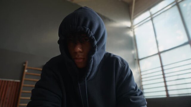 Tilt up of gloomy African American guy putting hood on his head and tying shoelaces of dark blue hoodie, crossing arms and putting them on knees while listening to psychologist at session