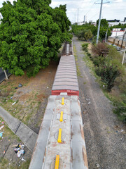 Vertical aerial drone shot showcasing a long freight train on railway tracks in Guadalajara, Mexico. Lush green trees line the urban route, with a glimpse of city infrastructure and a parallel road