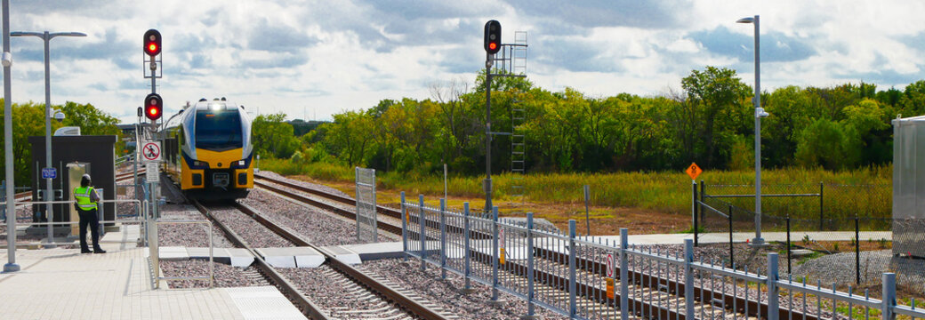 Panorama view electric light rail train approaches DFW station as staff member in safety vest monitors platform activity. Red signals and fencing ensure safe operations, lush green and open land
