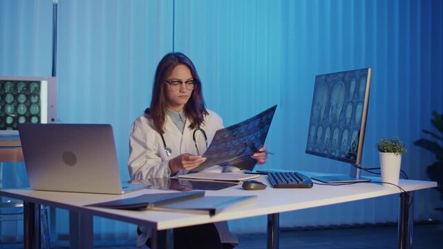 Female Doctor Studies MRI Scans on Desk and Computer Screen, Conducting Advanced Medical Diagnostics and Research. Concept of Innovation, Precision Healthcare, and Scientific Medical Analysis.
