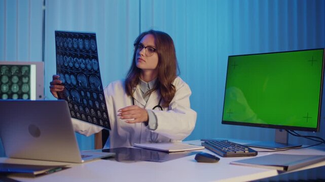 Female Doctor in White Coat Examines Medical Scans While Sitting at Desk With Laptop and Green Screen Monitor. Concept of Diagnostics, Digital Healthcare, and Medical Data Analysis for Research 