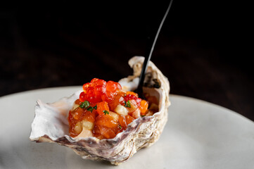 A close-up of a raw oyster on the half shell, topped with a tartare mix, bright orange salmon roe, and fresh herbs, served on a white plate with a fork