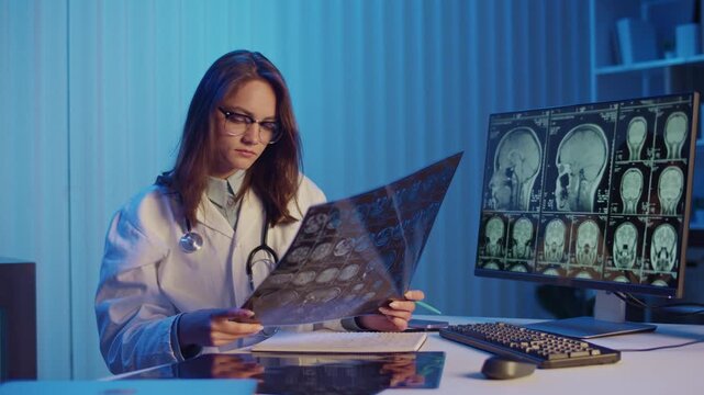 Female Doctor Studies MRI Scans on Desk and Computer Screen, Conducting Advanced Medical Diagnostics and Research. Concept of Innovation, Precision Healthcare, and Scientific Medical Analysis.