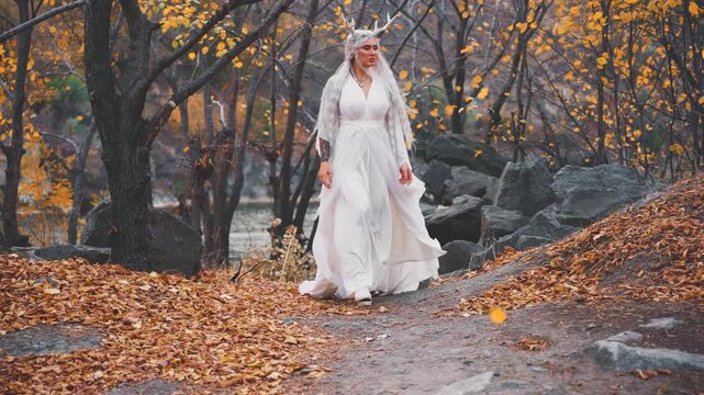 fantasy young woman faun walking in autumn forest enjoy yellow trees wind blow. sexy fairy tale girl long blonde hair, waving silk white dress slow motion. snow queen style crown deer antlers on head