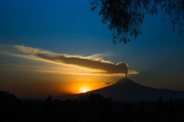 Mystical silhouette of Popocatépetl volcano erupting at sunrise. Fumarole over the peak, framed by tree branches
