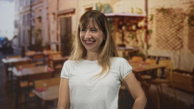 Woman smiling with hand on hip and bare arms at restaurant terrace outdoors, wearing white t shirt and laughing openly; happiness ease.
