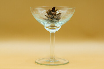 A clear, etched champagne glass, filled with clear liquid and containing a pinecone, is the central element against a muted, golden-yellow backdrop