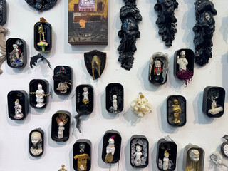 A wall installation featuring several small, dark-framed, shadow box-like enclosures containing miniature, doll-like ceramic figures and mixed media elements on a white surface.