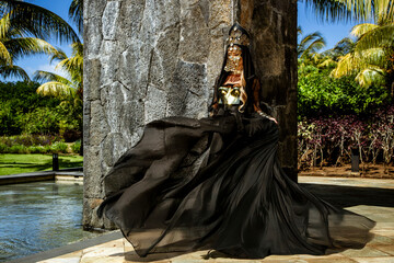 Mysterious woman in dramatic flowing black gown with gold mask posing by stone wall and tropical...