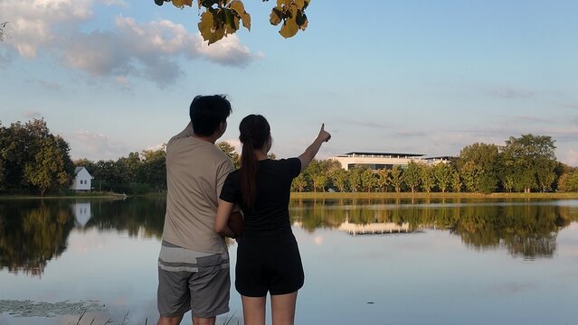 A couple is standing by a lake, pointing at a building in the distance