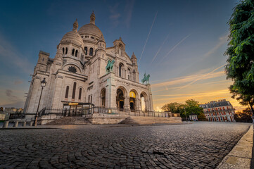 Fototapeta premium Visitors admire the stunning Sacre Coeur Basilica atop Montmartre as the sun sets, casting a warm glow on the iconic white domes and surrounding greenery under a blue sky.